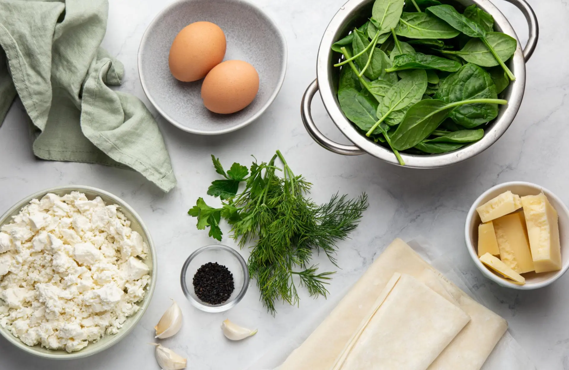 Fresh spanakopita ingredients on a marble surface: spinach in a colander, dill, parsley, crumbled Greek feta, eggs, phyllo pastry, and butter